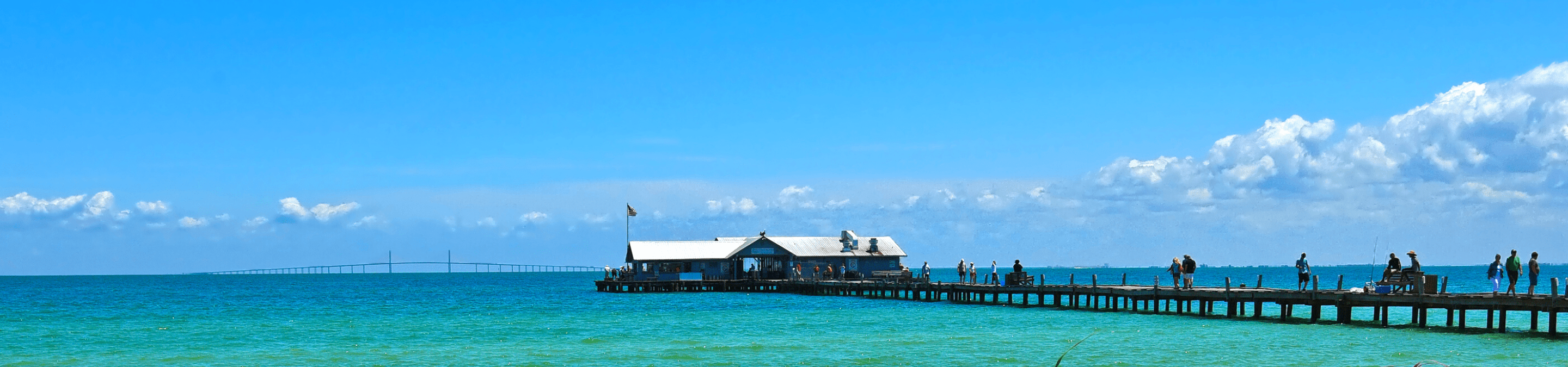 view of Anna Maria City Pier from the shore on a bright sunny day with the Skyway bridge in the background and people walking along the pier