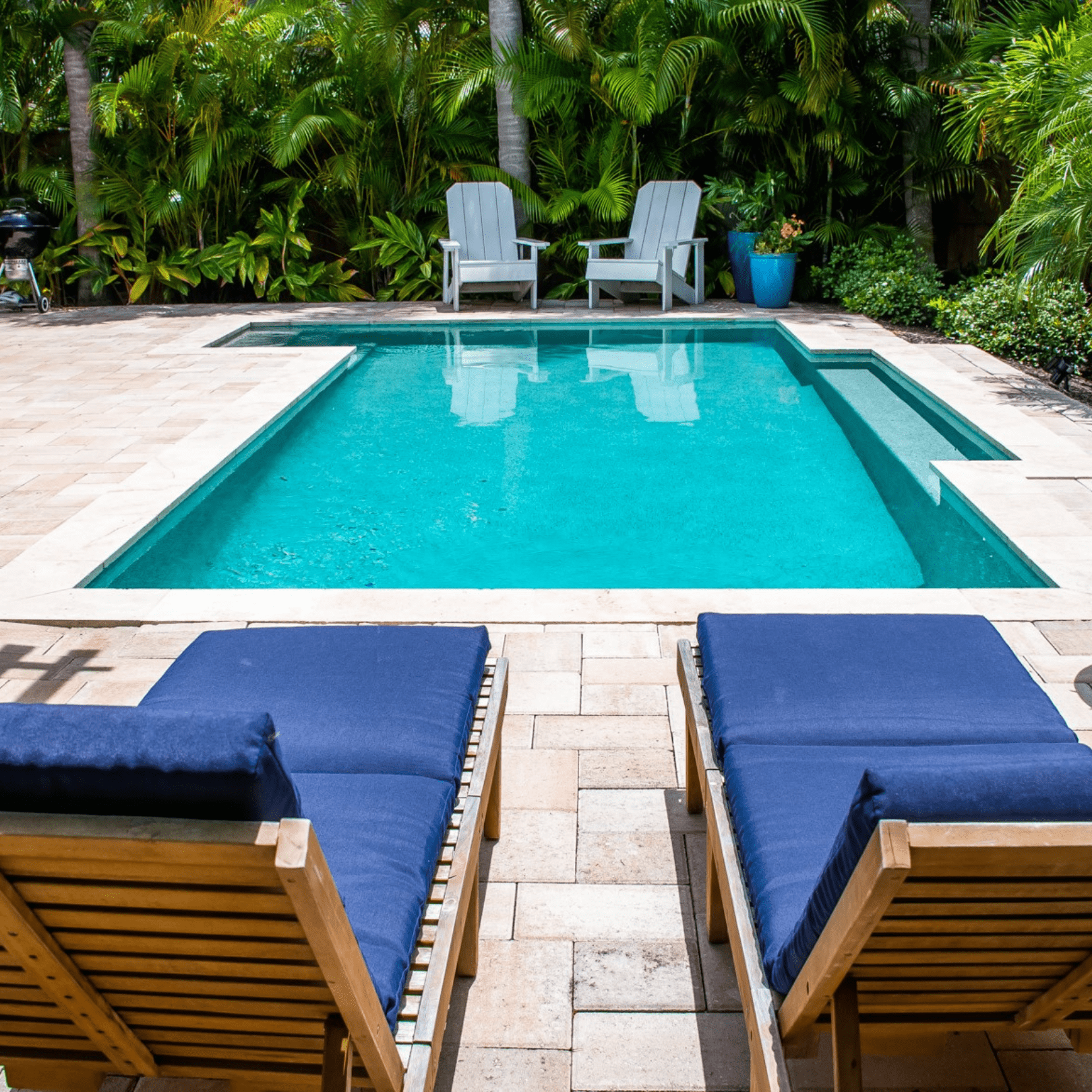view looking at a rectangular pool from behind two wooden loungers with royal blue cushions and two gray adirondack chairs at the other end of the pool of Island Time on Anna Maria Island