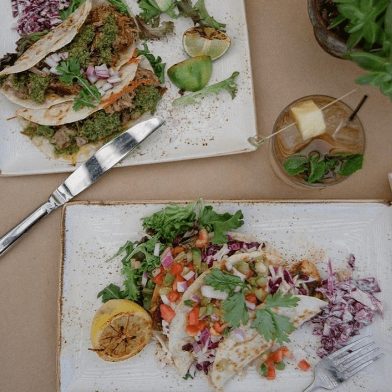 looking down at two rectangular plates of two tacos & a half lemon and hard drink between the two plates