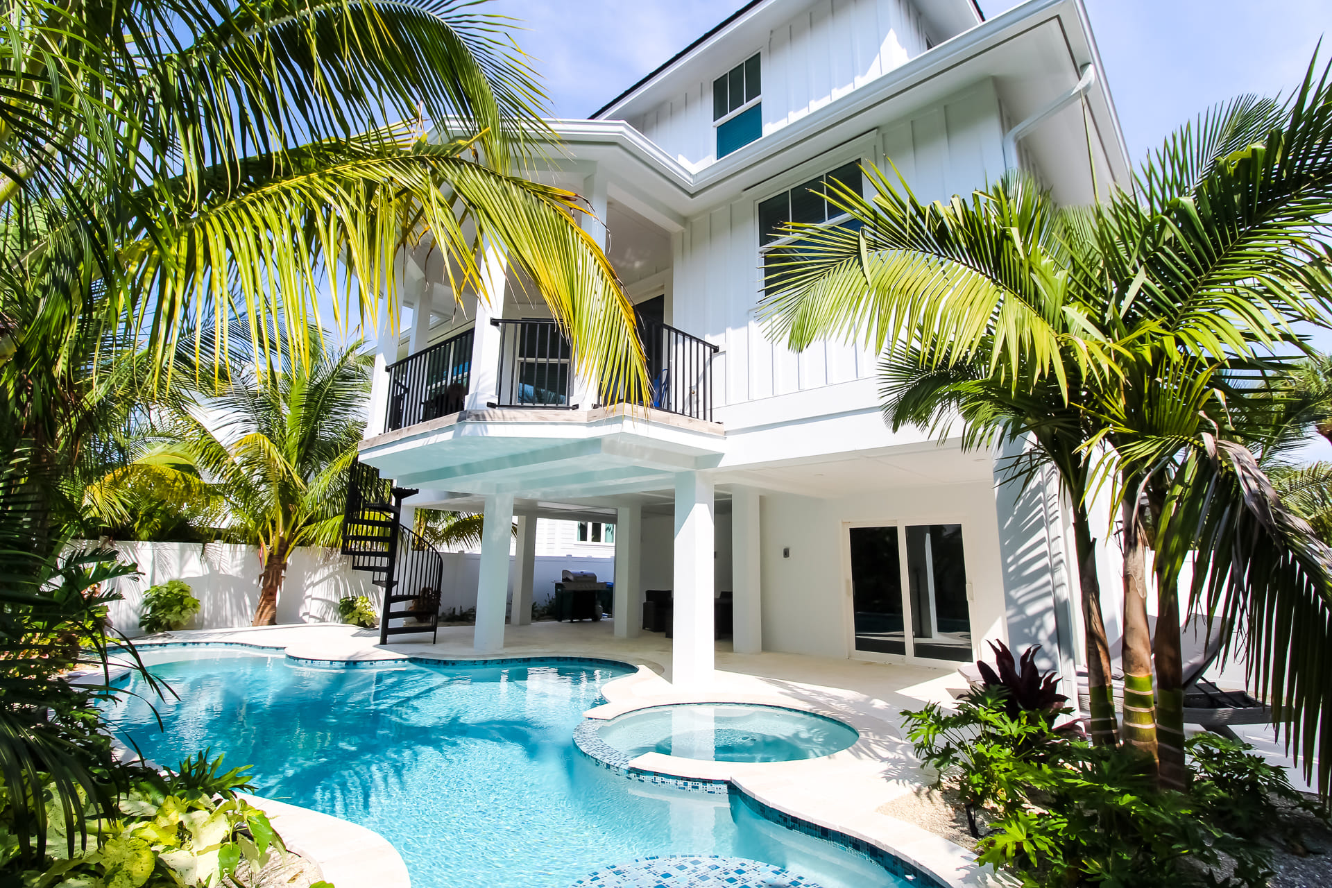 view from pool looking at a white three story house in full sun with spiral staircase leading to second floor, large pool & circular spa surrounded by foliage located on Anna Maria Island
