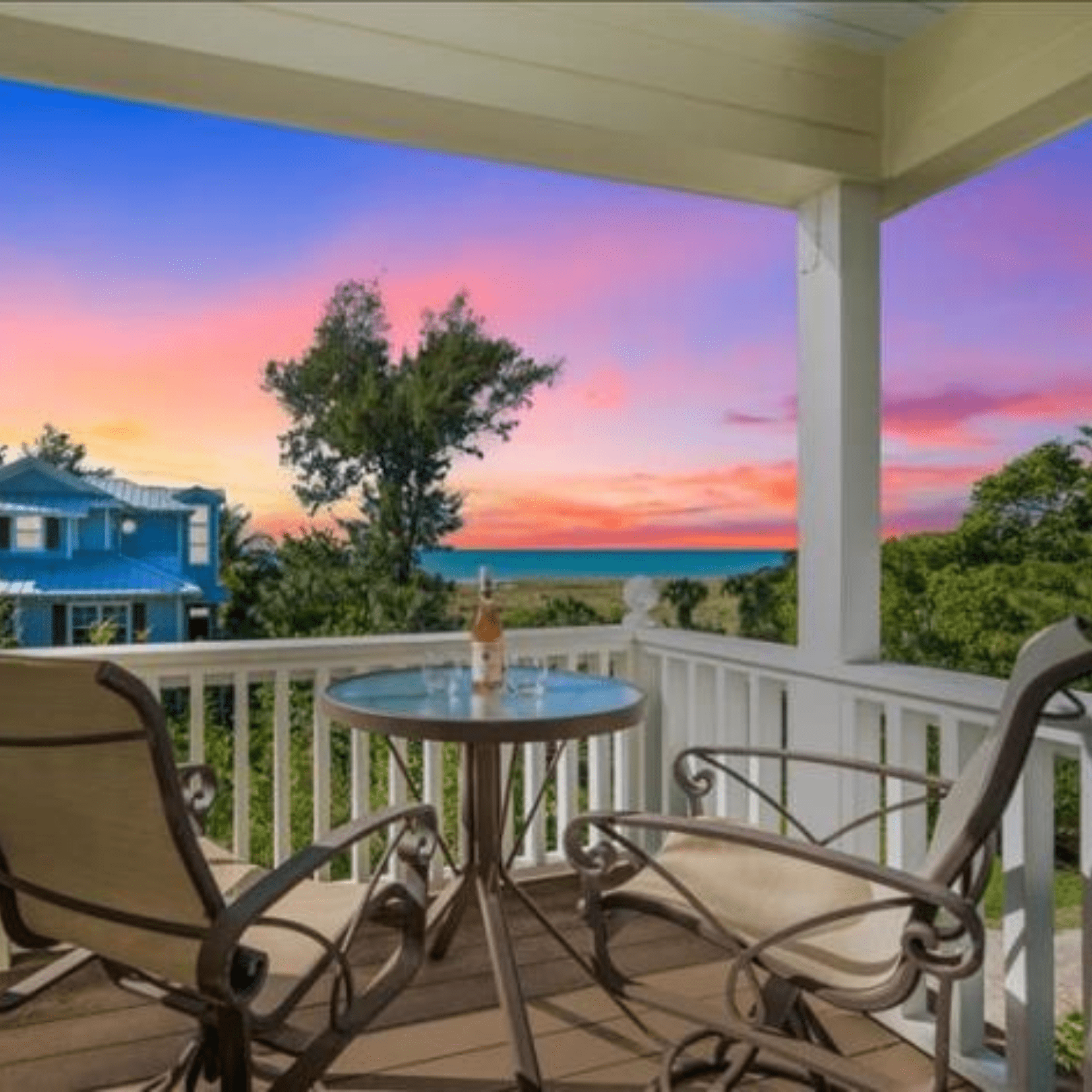 At our Anna Maria island vacation rentals here is a covered balcony view from behind two patio chairs and a small side table with a bottle of wine and two glasses looking out at a sunset off the coast of Anna Maria Island