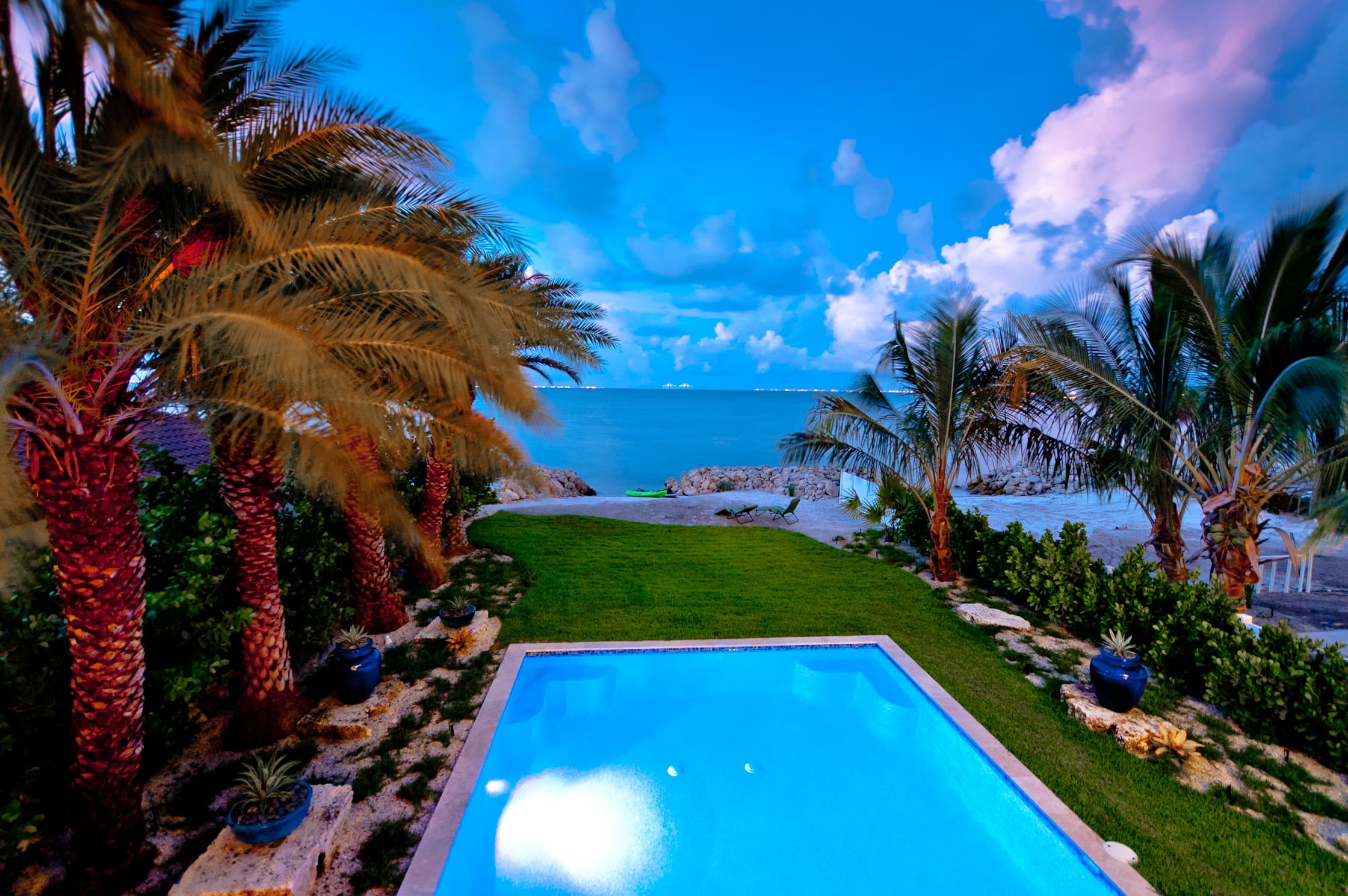 balcony view overlooking rectangular pool with palm trees on the left side with a lawn past the pool leading to a small sandy beach and private access to the bay off of Anna Maria Island
