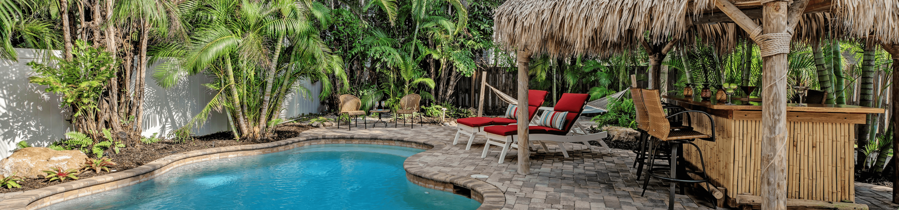 Our anna maria island 4 bedroom vacation rental with paver patio around circular pool surrounded by tall palm trees with a hammock in the background and a tiki hut with a bar to the right of the pool.