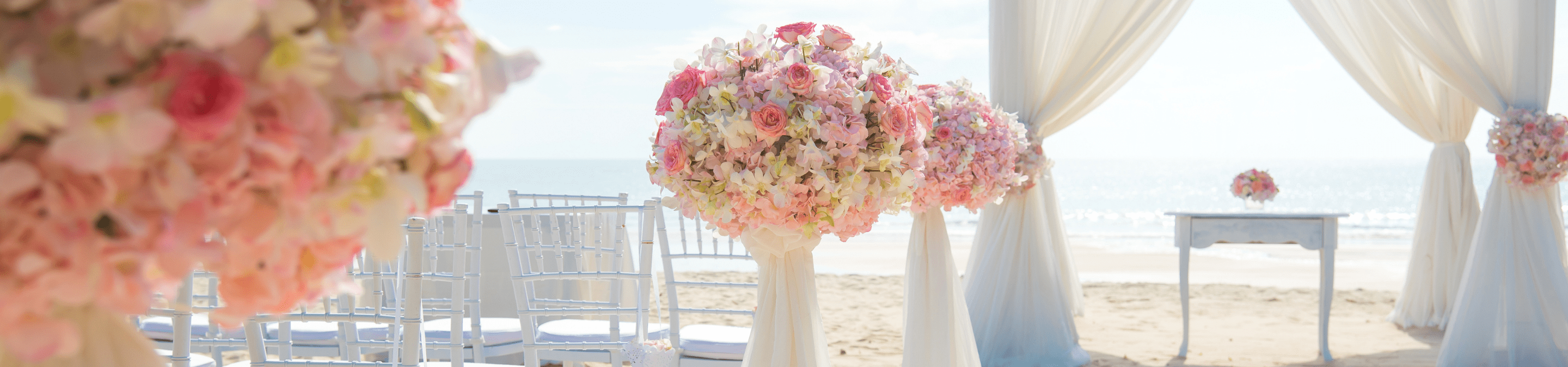 beach wedding with white bamboo chairs with large bundles of flowers lining the isle leading to a sheer curtain covered tend with small table in the middle of the text on a sunny day