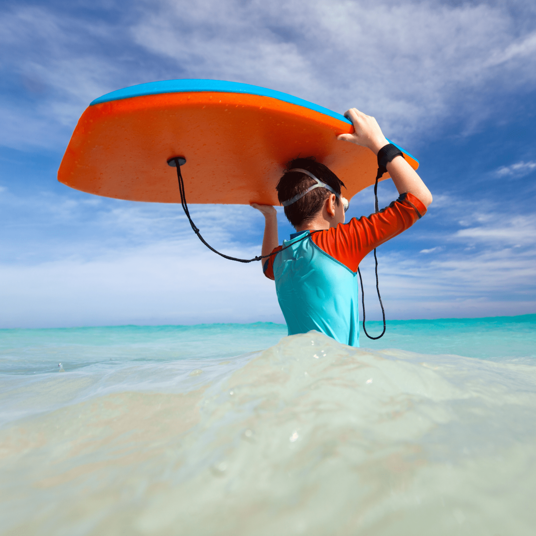 Boy caries a boogie board on his head as he wades into the ocean.