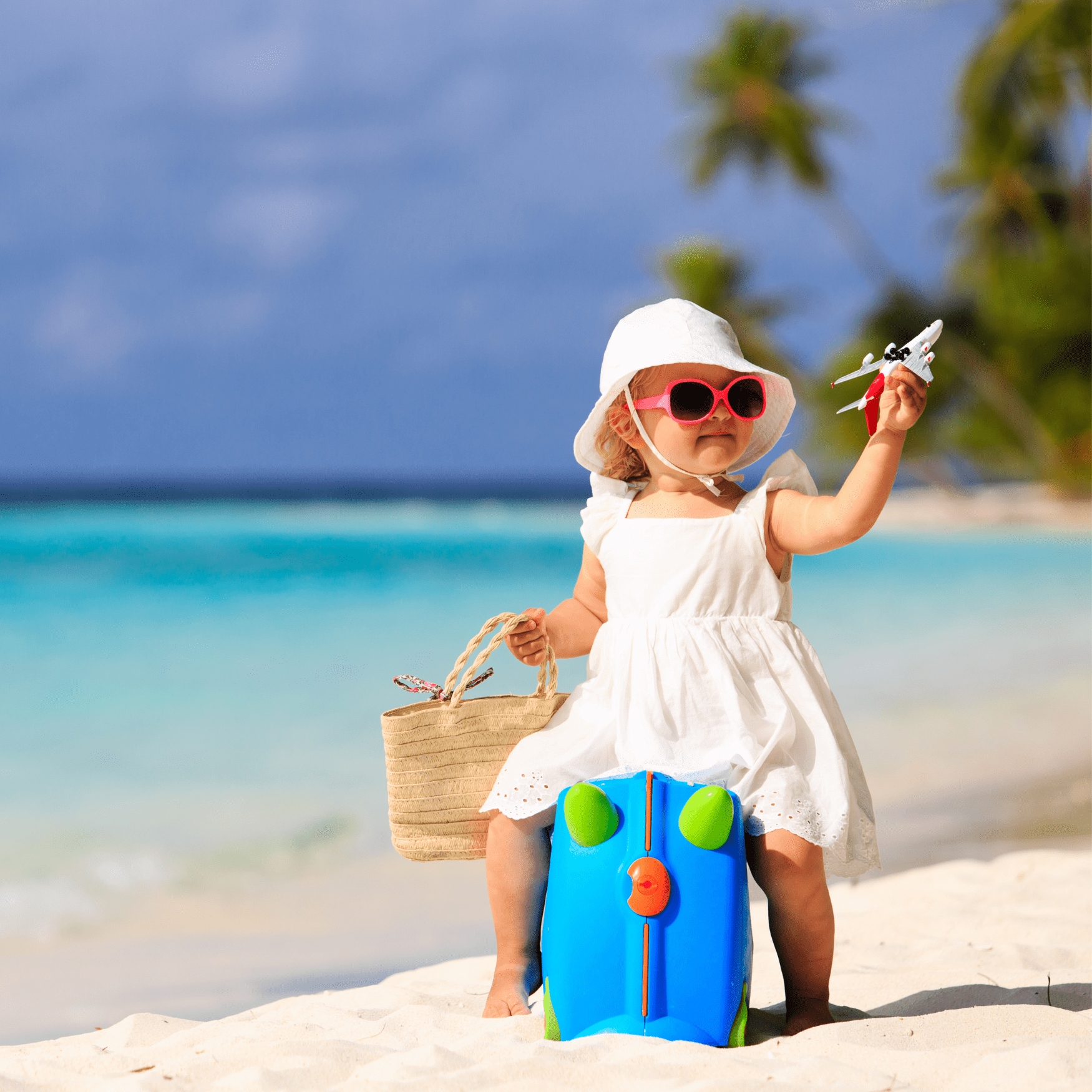 A baby sits on a suitcase on the beach, holding a bag in one hand and a toy airplane in the other.