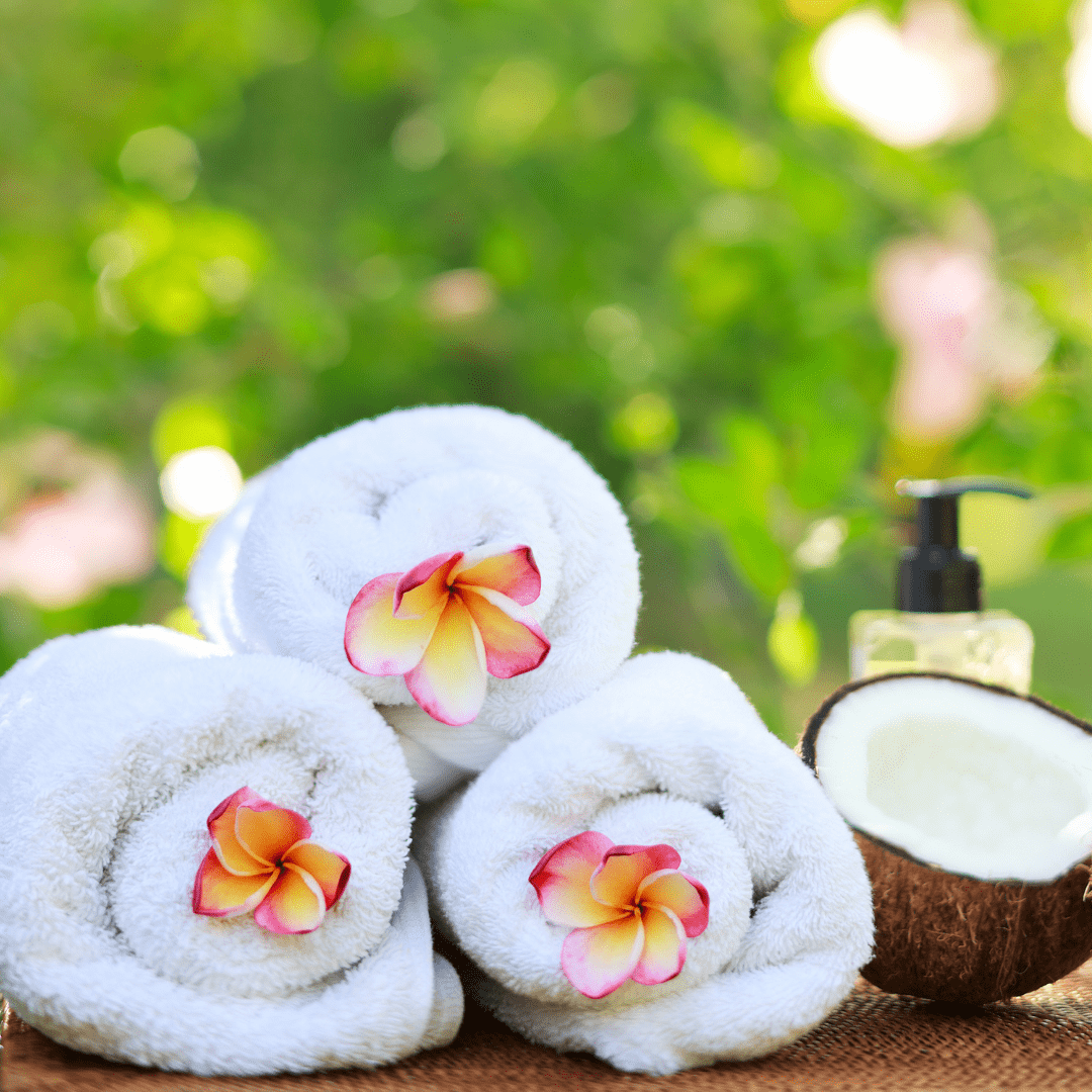 Three rolled towels with flowers on them, a half a coconut and a bottle of massage oil.