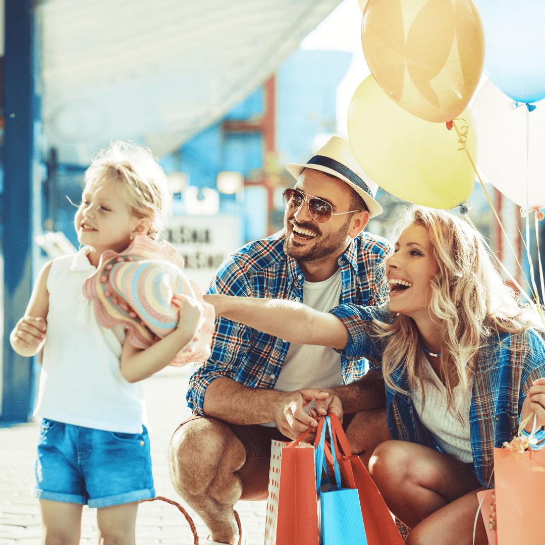 Couple and little girl with shopping bags and balloons pointing at something unseen.