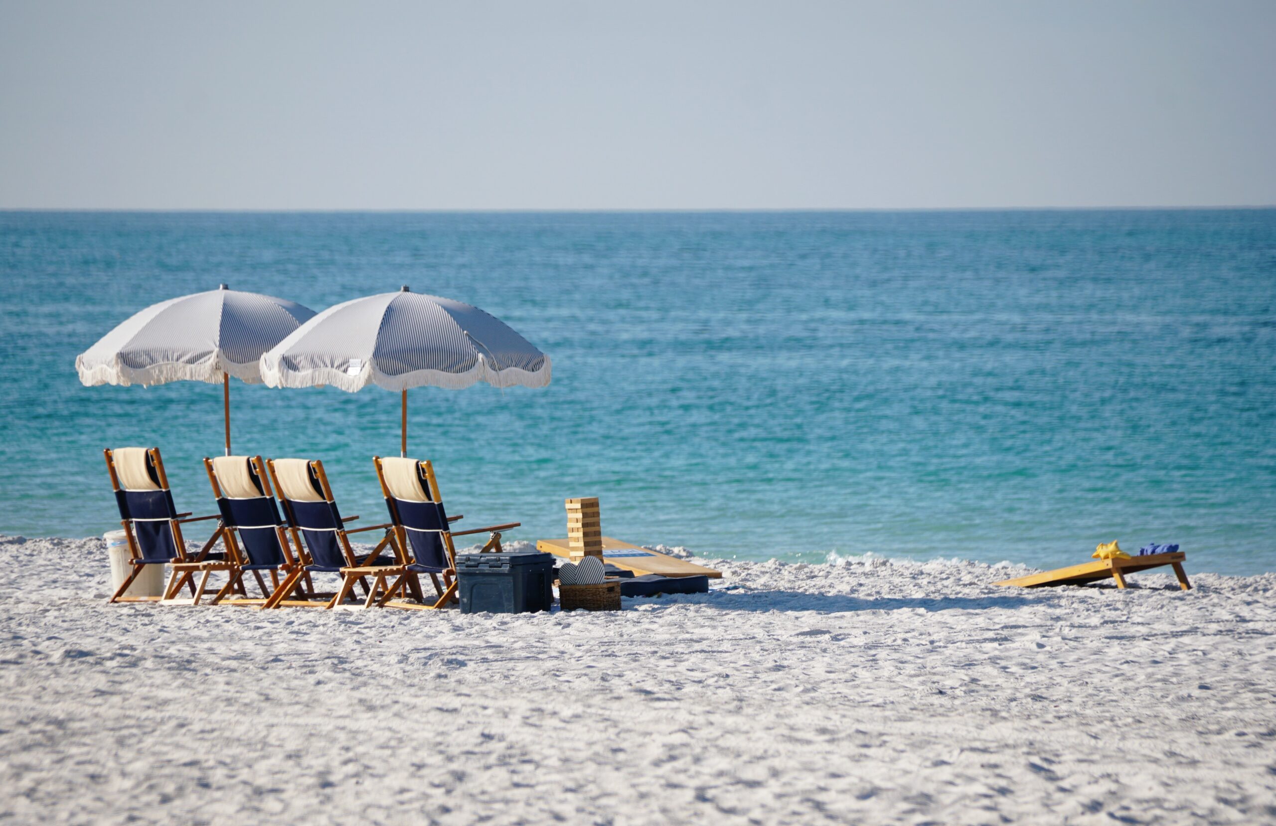 Four wooden lounge chairs under striped umbrellas facing calm turquoise water with a cooler, games and cornhole set near Palmetto Beach, Anna Maria Island, Florida, U.S.A