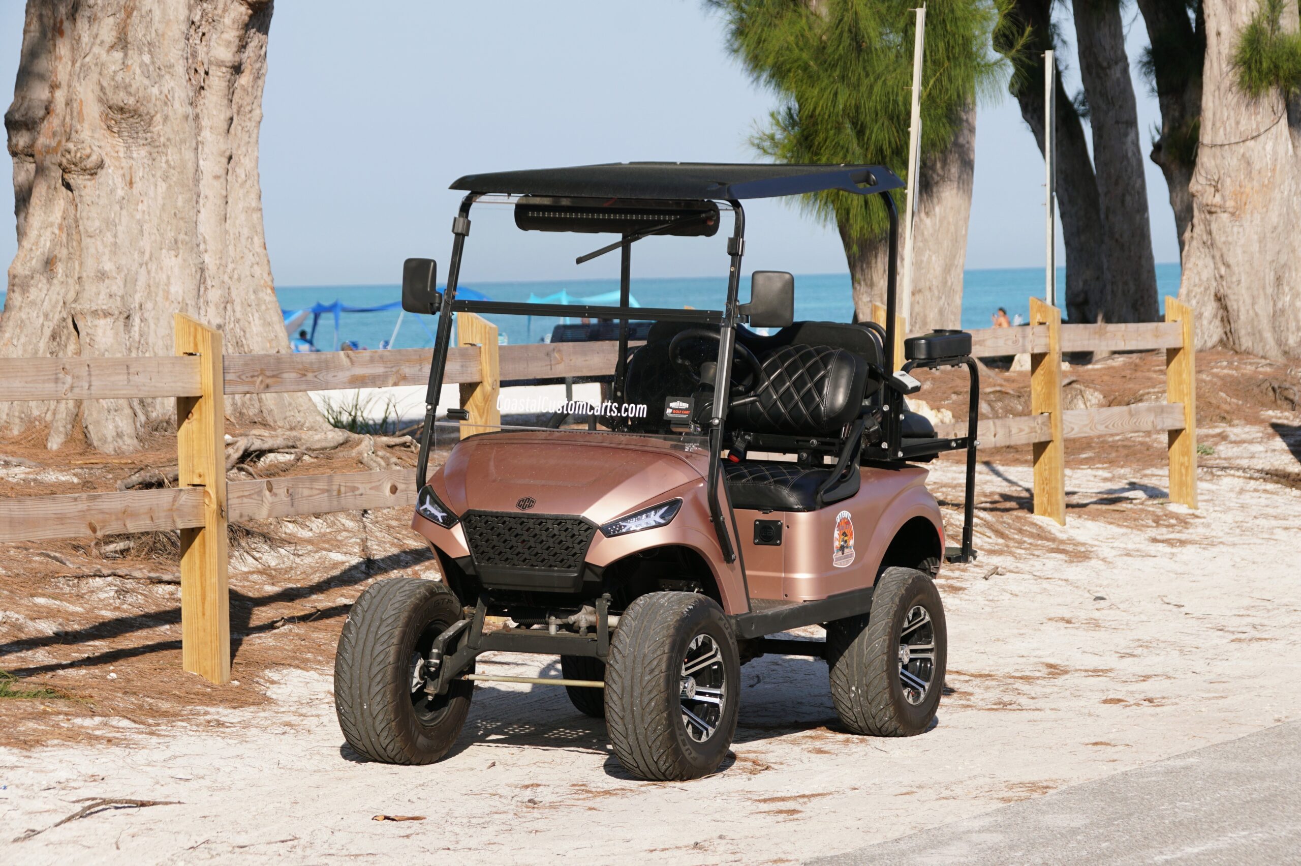 A bronze custom golf cart with off-road tires and stitched seats parked near a beachside path.